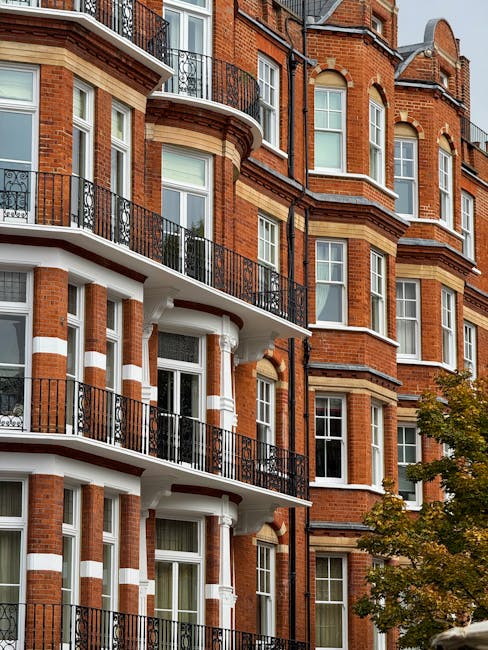 Exterior view of a historic red brick residential building in Lauderdale Mansions Maida Vale showcasing multiple floors with ornate white cornices and decorative brickwork. The building features large, white-framed sash windows, some with partially drawn curtains, and small balconies with black wrought iron railings. The photograph is taken during daytime with natural lighting, highlighting the clean and well-maintained facade. Surrounding trees with green foliage are visible in the foreground. This image relates to residential property access and maintenance, emphasizing the importance of surface cleaning and upkeep, as provided by Cleaners Maida Vale, experts in domestic cleaning and deep cleaning services for properties like Lauderdale Mansions Maida Vale.