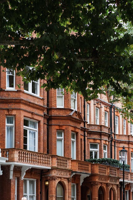 Exterior view of a historic red brick residential building in Lauderdale Mansions Maida Vale showcasing multiple floors with ornate white cornices and decorative brickwork. The building features large, white-framed sash windows, some with partially drawn curtains, and small balconies with black wrought iron railings. The photograph is taken during daytime with natural lighting, highlighting the clean and well-maintained facade. Surrounding trees with green foliage are visible in the foreground. This image relates to residential property access and maintenance, emphasizing the importance of surface cleaning and upkeep, as provided by Cleaners Maida Vale, experts in domestic cleaning and deep cleaning services for properties like Lauderdale Mansions Maida Vale.
