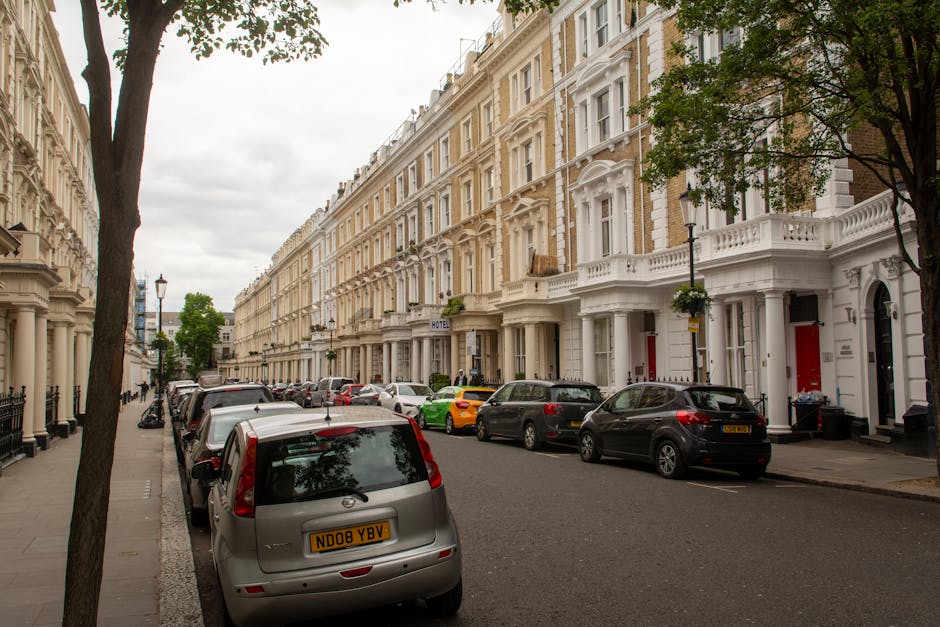 A residential street in Maida Vale W9 featuring a row of elegant Victorian-style terraced houses with white facades and decorative architectural details. The pavement is lined with parked cars, including silver, black, and yellow vehicles, with one silver car prominently in the foreground. Tall trees with green foliage provide shade along the sidewalk, and a lamppost is visible near the center of the image. The street appears clean and well-maintained, with soft natural lighting on a cloudy day, reflecting a tidy and tranquil neighbourhood typical of London. The image highlights the area where professional domestic cleaning services by Cleaners Maida Vale might operate, ensuring surfaces such as car exteriors and outdoor walkways are kept spotless and hygienic.