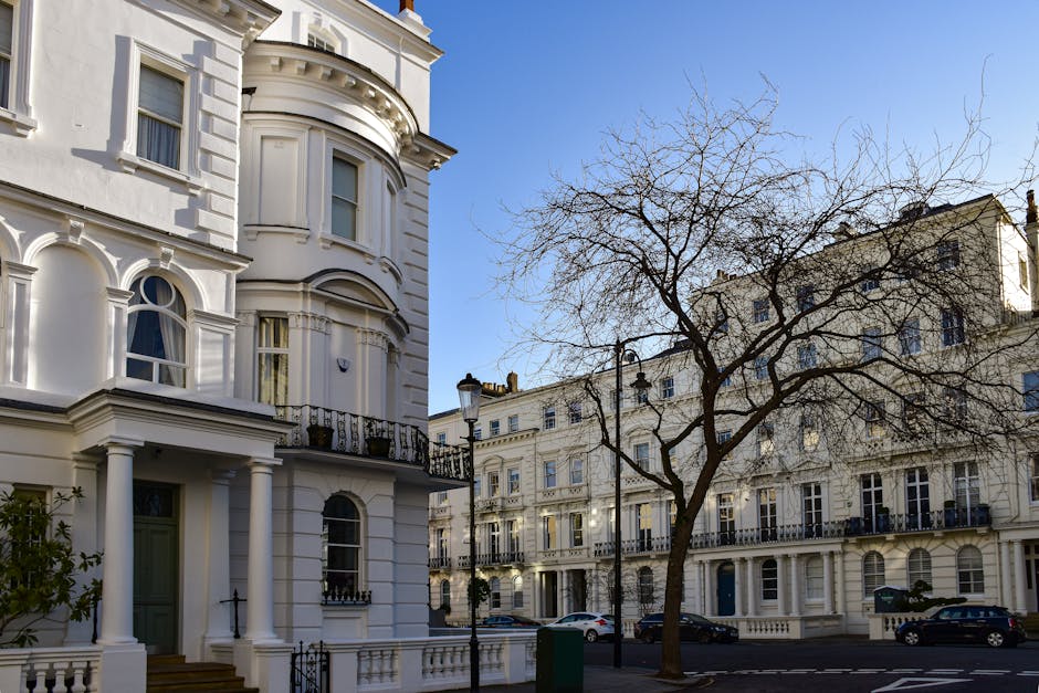 Photograph of a classic Victorian-style residential building with ornate white exterior features, including rounded bay windows, decorative cornices, and black wrought-iron balconies, set on a quiet street in Maida Vale. The building is surrounded by a paved sidewalk and a leafless tree in the foreground, with modern street lamps and parked cars lining the street under bright daylight. Clean, well-maintained surfaces reflect the sunlight, and the overall scene exudes a tidy and pristine appearance, characteristic of professional domestic cleaning or surface cleaning services provided by Cleaners Maida Vale. The image emphasizes the elegance and cleanliness of the residential area's architecture and external environment.