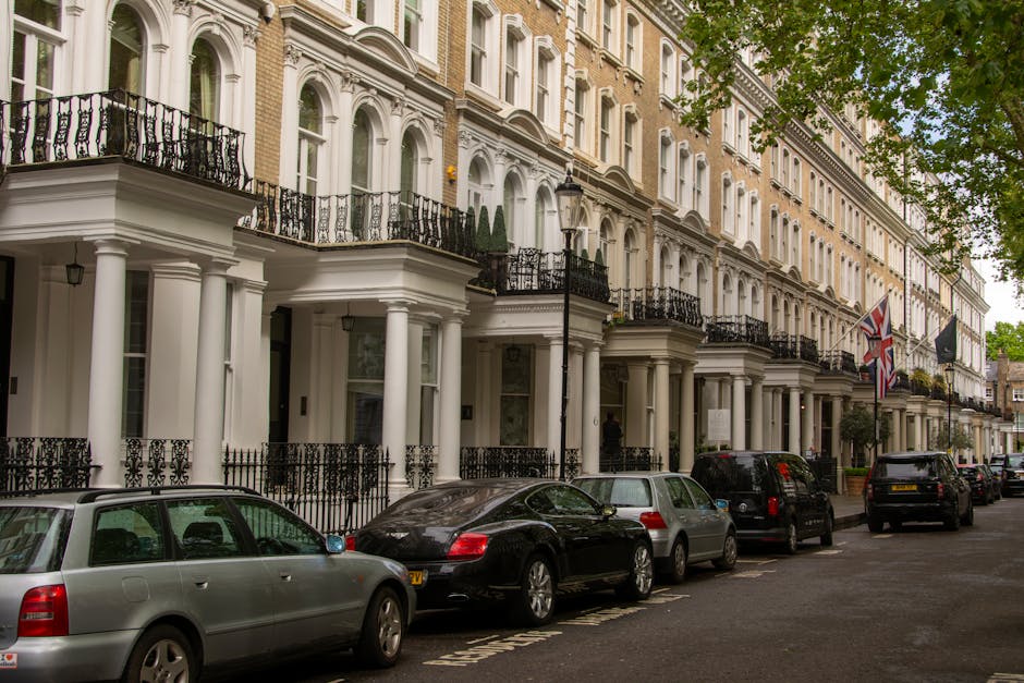 A residential street in Maida Vale W9 featuring a row of elegant Victorian-style terraced houses with white facades and decorative architectural details. The pavement is lined with parked cars, including silver, black, and yellow vehicles, with one silver car prominently in the foreground. Tall trees with green foliage provide shade along the sidewalk, and a lamppost is visible near the center of the image. The street appears clean and well-maintained, with soft natural lighting on a cloudy day, reflecting a tidy and tranquil neighbourhood typical of London. The image highlights the area where professional domestic cleaning services by Cleaners Maida Vale might operate, ensuring surfaces such as car exteriors and outdoor walkways are kept spotless and hygienic.