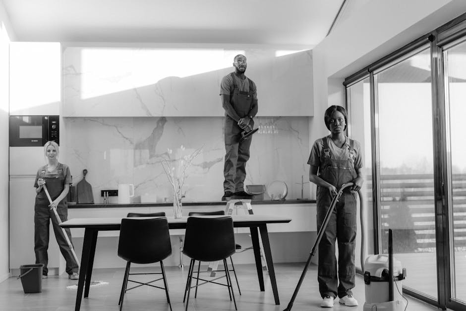 A black-and-white photo of three professional cleaners inside a modern, bright kitchen. Two women are standing on the floor, holding mops and cleaning tools, while a man stands on a small step ladder,
