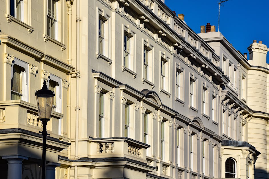 Two professional window cleaners are performing surface cleaning on the exterior glass facade of a commercial building in Maida Vale. One worker, dressed in a blue jacket and orange gloves, is suspend