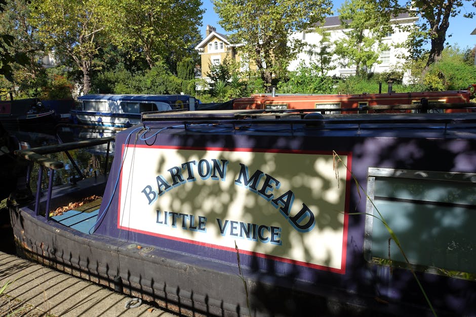 Close-up of a narrowboat moored along the Little Venice canal in Maida Vale, with the boat's white sign displaying 'Barton Mead Little Venice' in bold blue letters. The boat features a black and green painted surface, and its wooden deck and stainless steel railings are visible beneath the sign. The canal water is calm, reflecting the surrounding lush green trees and nearby residential houses with white facades and pitched roofs in the background. Bright daylight illuminates the scene, highlighting the clean and well-maintained appearance of the boat and the vibrant greenery of the trees, emphasizing the importance of surface cleaning and maintenance for canal boats as part of routine deep cleaning services offered by Cleaners Maida Vale.