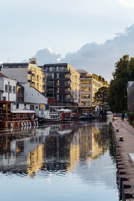 A scenic view of the Little Venice canal in Maida Vale featuring modern apartment buildings with glass and concrete facades alongside floating houseboats. The calm water reflects the structures and the partly cloudy sky above, with a paved walkway and greenery visible on the right. The scene exudes a clean, well-maintained appearance, highlighting the importance of surface cleaning and sanitisation in this residential area. For professional deep cleaning and domestic cleaning services in Maida Vale, residents can contact Cleaners Maida Vale to maintain the hygiene and shine of their properties.