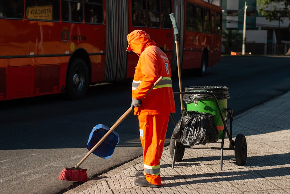 A sanitation worker wearing a beige cap, face mask, and gloves walks on a street while pulling a small blue trolley cart with an attached large orange waste bin and a smaller brown bin. The background