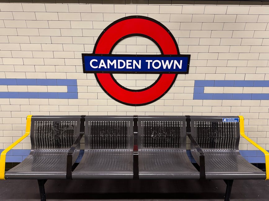 A row of four metal seating benches with perforated surfaces and black frames, located against a beige tiled wall at Camden Town underground station. The benches have yellow armrests on both ends, and a small blue and white safety sign is affixed to the right side. The background features large London Underground signage with a red circle, a blue bar, and the words 'Camden Town' in white letters. The seating area appears clean and well-maintained under bright lighting, with the tiled wall and station floor also looking tidy, reflecting professional cleaning and hygiene standards essential for a safe public environment. This image highlights the importance of surface cleaning and sanitisation in high-traffic transport hubs, exemplified by the vigilant maintenance carried out by [COMPANY_NAME].