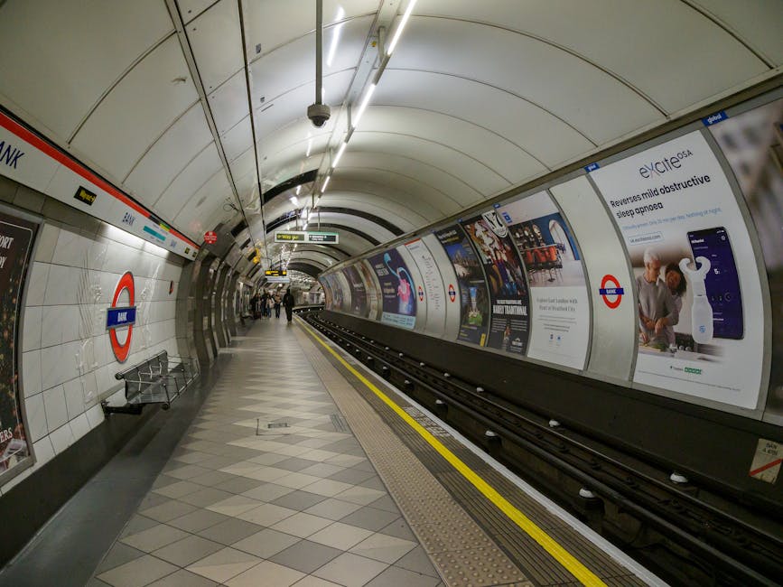 The image depicts an underground train station platform in Maida Vale, with a curved, tiled ceiling illuminated by fluorescent and LED lighting. The platform surface is clean and polished, with a section of tactile paving near the edge for safety. On the left side, there are metal benches and a few passengers seated, wearing masks. The walls feature advertising posters and signage, including symbols for the London Underground and station information. Tracks extend into a dark tunnel at the center of the image, suggesting a deep, underground setting. The overall scene is well-maintained, emphasizing hygiene and cleanliness in an urban public transportation environment, consistent with the professional cleaning and sanitisation services provided by Cleaners Maida Vale, as referenced in the page titled 'Maida Vale tube insider cleaning tips Randolph Avenue, Maida Vale'.