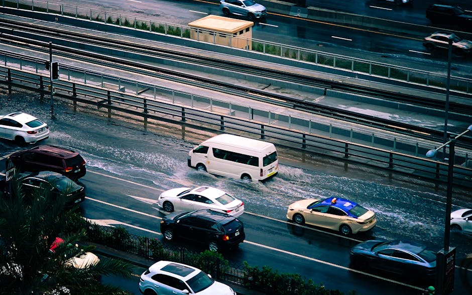 A busy multi-lane road in Maida Vale with several cars, including a white van and a beige taxi, driving through a floodwater that has accumulated across the street. The wet asphalt reflects the vehicl