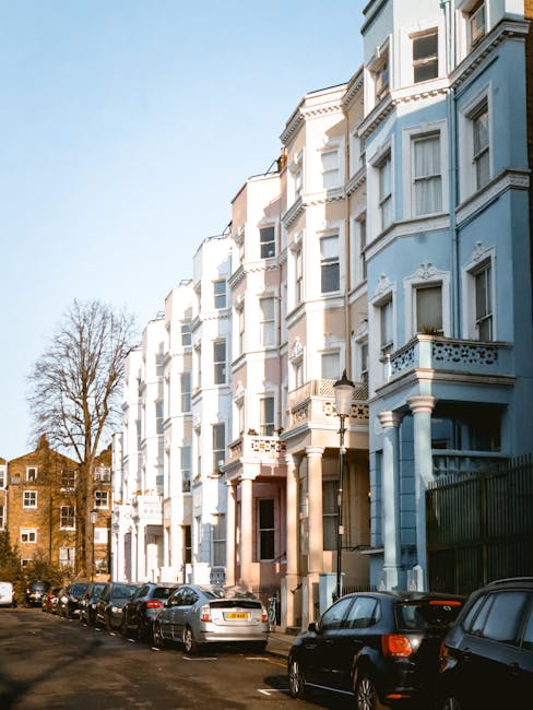 Exterior view of a row of white and light blue Victorian-style residential flats with decorative architectural details on the facades. The buildings feature bay windows, balconies with ornate railings, and classical columns at the entrance. Parking spaces along the street are occupied by various cars, and leafless trees are visible in the background under a clear blue sky. The scene is well-lit by natural sunlight, highlighting the clean, well-maintained appearance of the buildings. This setting reflects the type of residential environment where professional cleaning services, such as those provided by Cleaners Maida Vale, can perform surface cleaning, deep cleaning, and sanitisation to maintain hygiene and aesthetic appeal in flats like those on Elgin Avenue in Maida Vale.