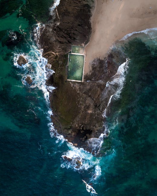 An aerial view of a small coastal area showing a sandy beach and a rocky outcrop with a rectangular pool filled with green water, situated on top of the rocks near the shoreline. The surrounding ocean has varying shades of blue and turquoise with visible waves crashing against the rocks. The scene is illuminated by natural sunlight, highlighting the textures of the rocks, sand, and water. This coastal landscape emphasizes natural beauty alongside man-made elements, suitable for content related to outdoor or leisure cleaning or maintenance by Cleaners Maida Vale, based on the context of their services for commercial cleaning at Warwick Avenue shops in Maida Vale.