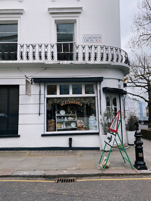 The ground floor of a white building with a curved black railing on the upper balcony, displaying a sign for Clarendon Cross NW1. The shop has a large front window showcasing baked goods and kitchenware, with a black awning above. Outside, a green and red A-frame sidewalk sign and a black bollard are visible on the grey pavement. The scene is well-lit with natural daylight, and the exterior appears clean and well-maintained, reflecting professional surface cleaning and maintenance by Cleaners Maida Vale for commercial and shopfront hygiene.
