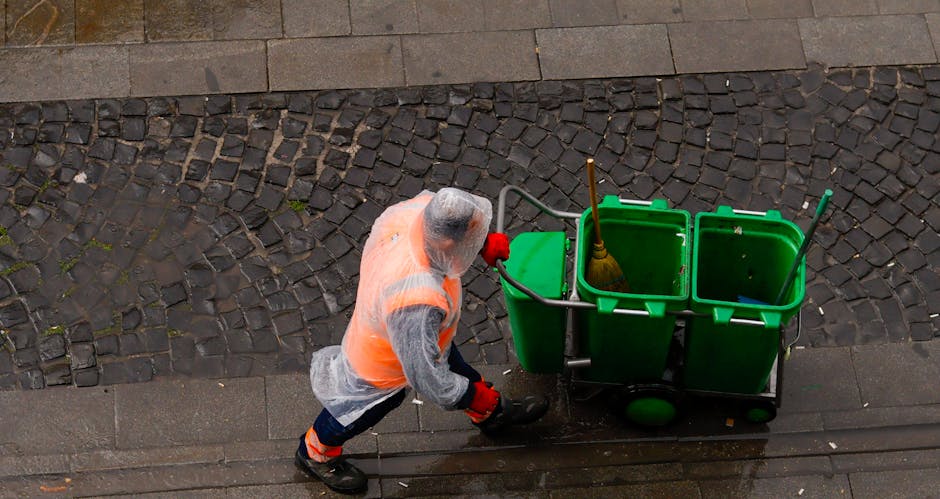 A cleaning professional from Cleaners Maida Vale is operating a green commercial cleaning cart containing three connected bins — one holding a mop with a yellow head, and the others for waste collection — on a paved sidewalk adjacent to a cobblestone street. The worker is dressed in a high-visibility orange vest, gloves, and a transparent raincoat, suggesting readiness for outdoor sanitation tasks in an urban area. The surface being cleaned features wet, dark-toned tiles and cobblestones, indicating ongoing or recently completed cleaning activities that contribute to hygiene maintenance consistent with Westminster Council waste rules in Maida Vale. Bright natural lighting highlights the cleanliness of the area, reflecting the professional approach to surface cleaning, deep cleaning, and sanitation that the company specializes in.