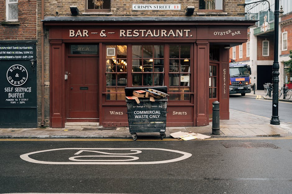 A view of a small restaurant with a red wooden facade and large glass windows, situated on a street corner under a street sign reading 'Crispin Street E1'. In front of the restaurant, there is a black waste bin labeled 'Commercial Waste Only' filled with cardboard boxes and paper debris, with some discarded papers lying on the sidewalk nearby. The establishment advertises wines, spirits, and cocktails on the window and side panel. The street features a double yellow line and a painted white symbol on the asphalt indicating a no-parking zone. To the right, a black bollard and a lamppost are visible, along with parked bicycles and a delivery truck in the background. The scene is captured in natural daylight, emphasizing the clean, well-maintained exterior of the property, typical of urban commercial premises. This image exemplifies the importance of proper waste disposal and surface cleanliness for businesses like those managed by Cleaners Maida Vale, following Westminster Council waste regulations for Maida Vale.