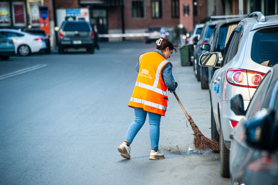 A female street cleaner wearing an orange high-visibility vest with the Cleaners Maida Vale logo is sweeping debris from the asphalt near parked cars on a residential street. She is using a traditional broom with a wooden handle and natural bristles, focusing on cleaning the curbside area. The street is lined with various cars, including a silver hatchback and a white sedan, parked parallel to the sidewalk. In the background, there are additional parked vehicles and a building with large windows, illuminated by natural daylight. The scene emphasizes surface cleaning and maintenance activities associated with ensuring hygienic, well-maintained parking zones in Maida Vale, reflecting professional cleaning standards.