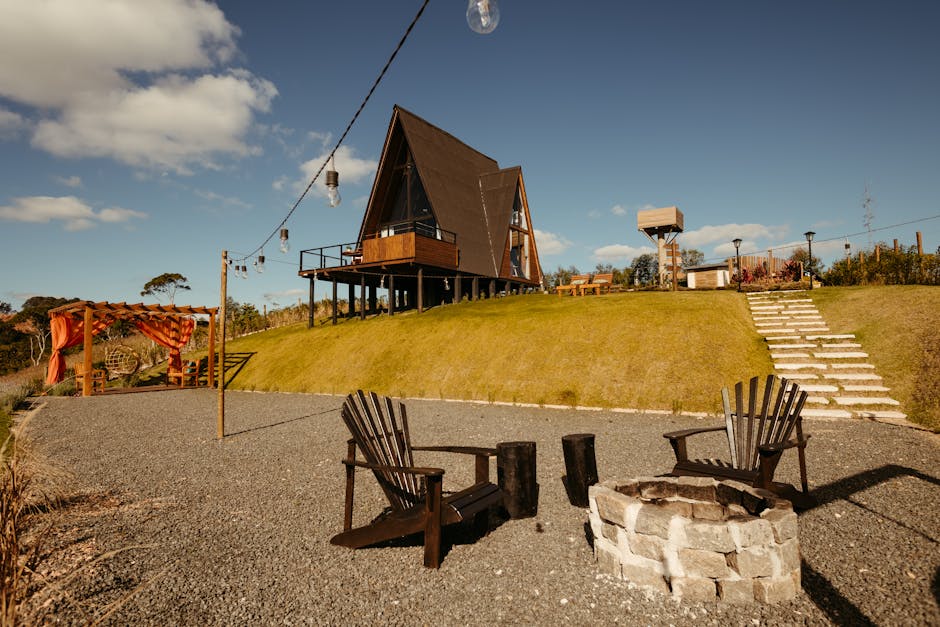 A spacious outdoor area featuring a modern, triangular wooden house elevated on stilts on a grassy hill under a bright blue sky with scattered clouds. In the foreground, two dark wooden Adirondack chairs frame a small round fire pit made of light-colored bricks, situated on a gravel surface. To the left, a wooden pergola with orange curtains provides shaded seating, and a string of outdoor lights is suspended overhead across the space. Stone steps lead up the grassy slope towards the house, with a wooden fence and a birdhouse visible in the background. The area appears well-maintained, clean, and designed for outdoor relaxation, reflecting high standards of cleanliness and outdoor surface maintenance typical of professional cleaning services like those from Cleaners Maida Vale.